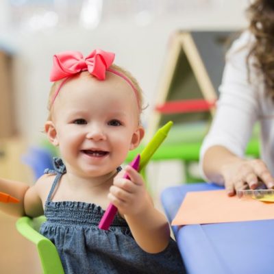 Cute little toddler girl sitting at a table and playing with crayons with her teacher in the background. She is holding the crayons and smiling at the camera.